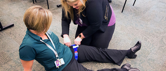 Female class attendee in a seated position on the floor, while female trainer demonstrates proper placement of a leg tourniquet