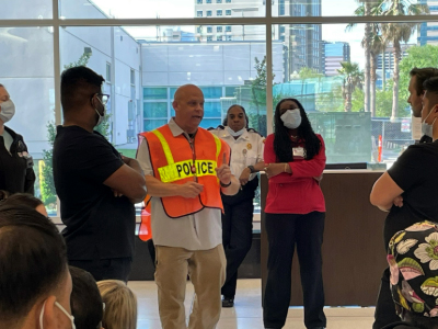 Candid image of a group of people gathered around an officer who is speaking to everyone and wearing a bright orange 'Police' safety vest.